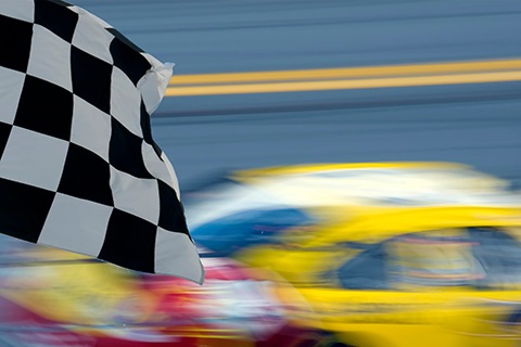 A checkered flag in front of a race car speeding by on a track.